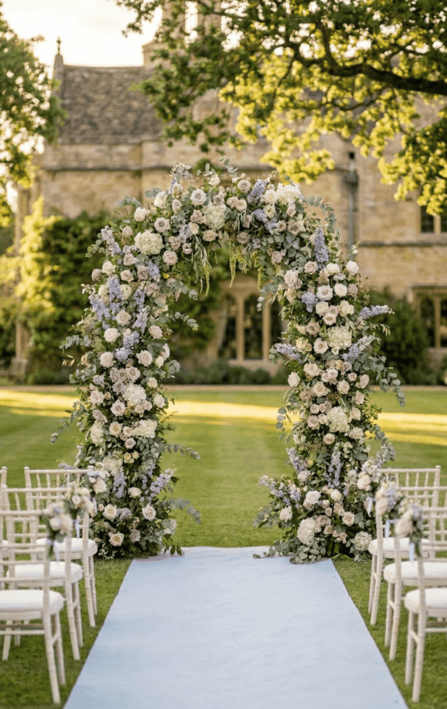 A romantic outdoor spring garden wedding ceremony setup featuring a lush floral arch over a stone altar, with white chairs, a winding pathway, and scenic green hills in the soft natural sunlight.